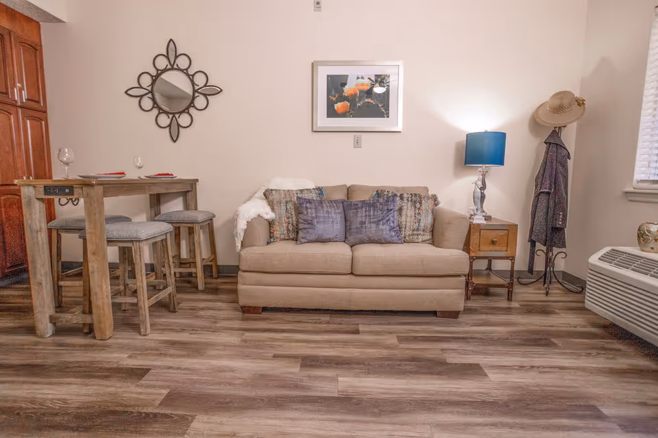 A cozy living room area featuring a beige sofa with multiple decorative pillows and a white throw blanket. To the left, there is a wooden high dining table with four cushioned stools and two wine glasses on top. On the wall above the sofa, there is a framed picture of orange flowers and a decorative round mirror. To the right of the sofa, there is a wooden side table with a blue lamp and a coat rack holding a hat and a jacket. The room has wood flooring and a window with blinds on the right side.
