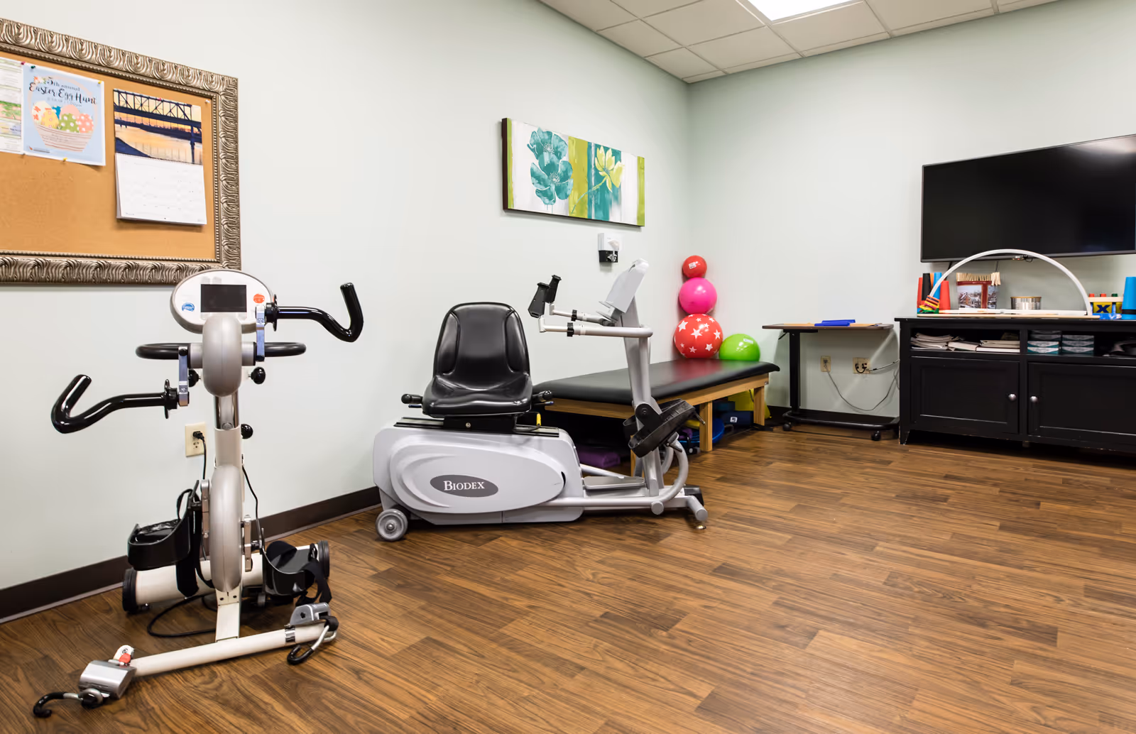 Small rehabilitation room with two exercise machines, a treatment table, colorful therapy balls, wall art and a TV cabinet.