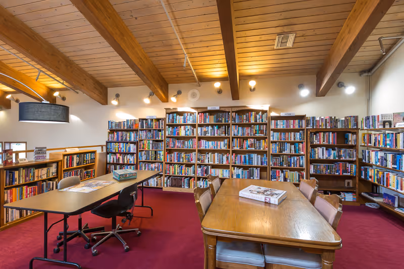 Well-lit library/reading room with bookshelves along the walls, tables and chairs on red carpet under exposed wooden beams.