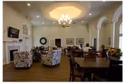 A spacious senior living common room with dining tables and patterned armchairs beneath a chandelier-lit ceiling and a fireplace, with several residents seated.