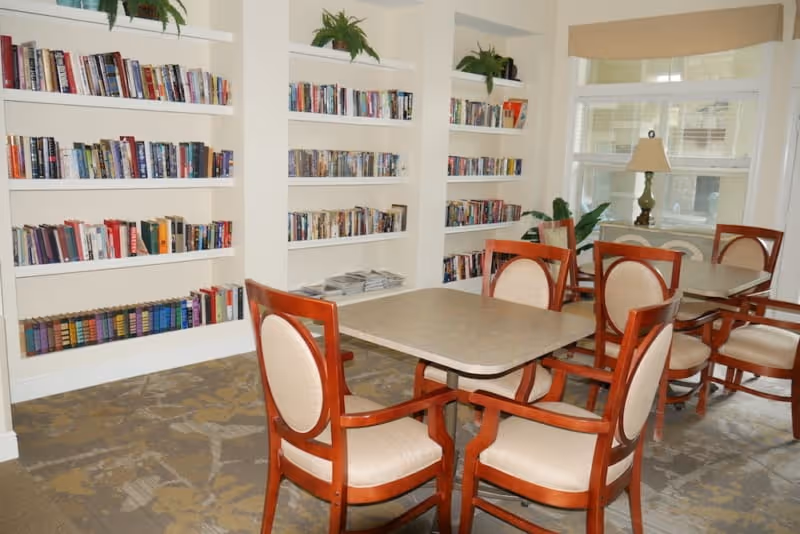 A cozy reading and seating area in a senior living facility with wooden chairs upholstered in beige fabric around square tables. The walls feature built-in white bookshelves filled with books and some potted plants on top. A window with a beige valance lets in natural light, and a table lamp is placed on a small table near the window.