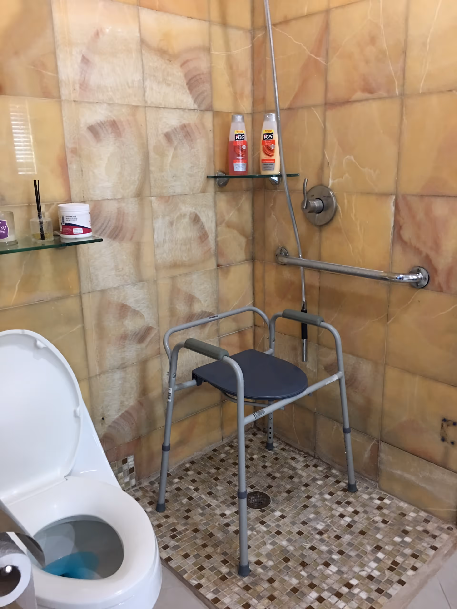 Shower area with a portable shower chair and grab bar next to a toilet in a tiled bathroom.