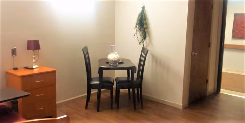Small dining area in a nursing facility with a square table and four chairs against a wall, a dresser with a lamp, and a doorway to a hallway.