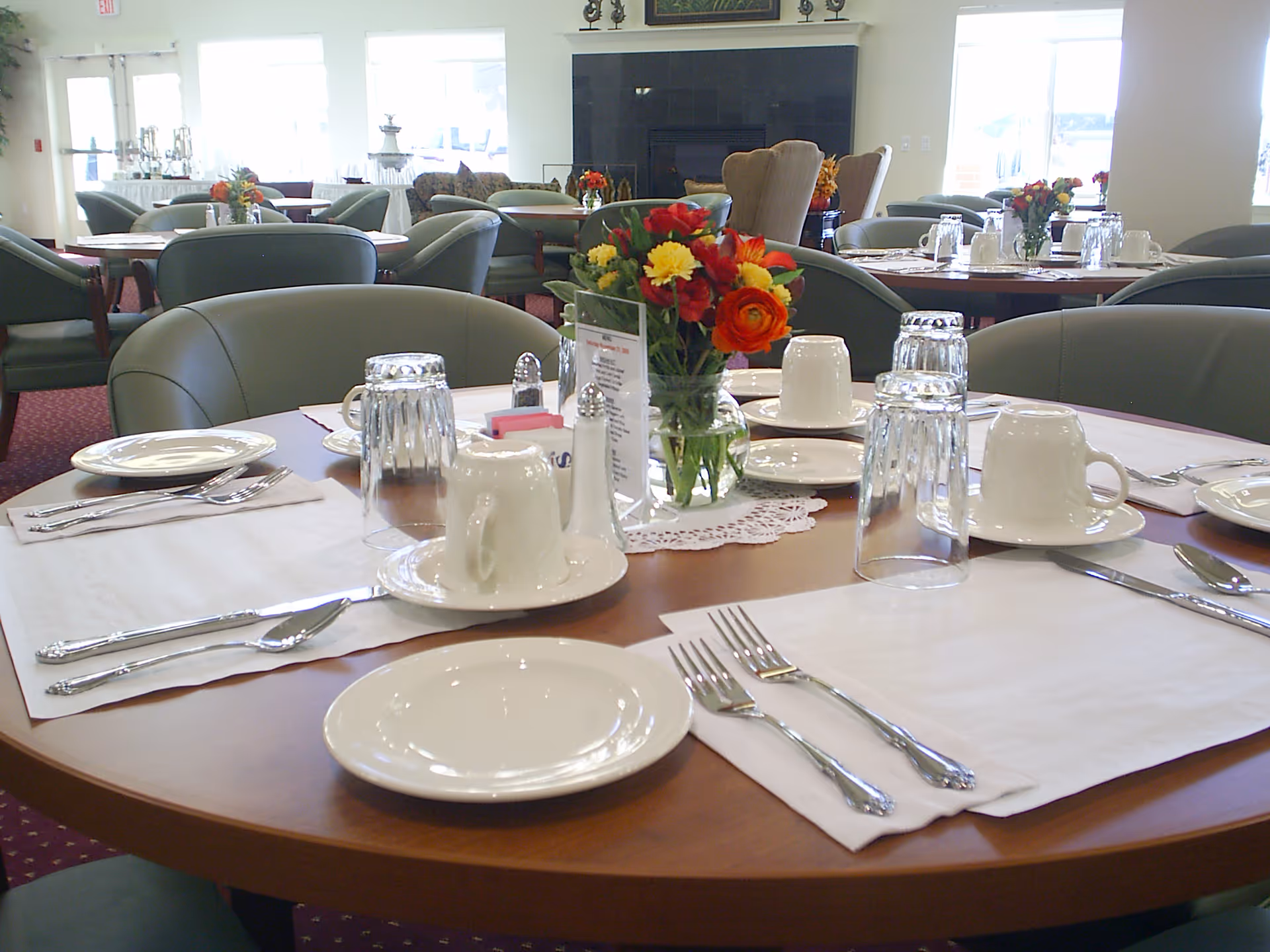A dining room table set for a meal with white plates, cups, silverware, and upside-down glasses. A small vase with colorful flowers is placed in the center of the table. In the background, there are more tables and chairs, large windows letting in natural light, and a fireplace with armchairs nearby.
