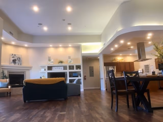 Spacious open-concept living area in a senior living facility featuring a seating area with a dark green couch and a yellow throw, a fireplace on the left wall, built-in shelves with decorative items, and a kitchen with wooden cabinets and a dining table with chairs on the right side. The room has recessed lighting and wood flooring.