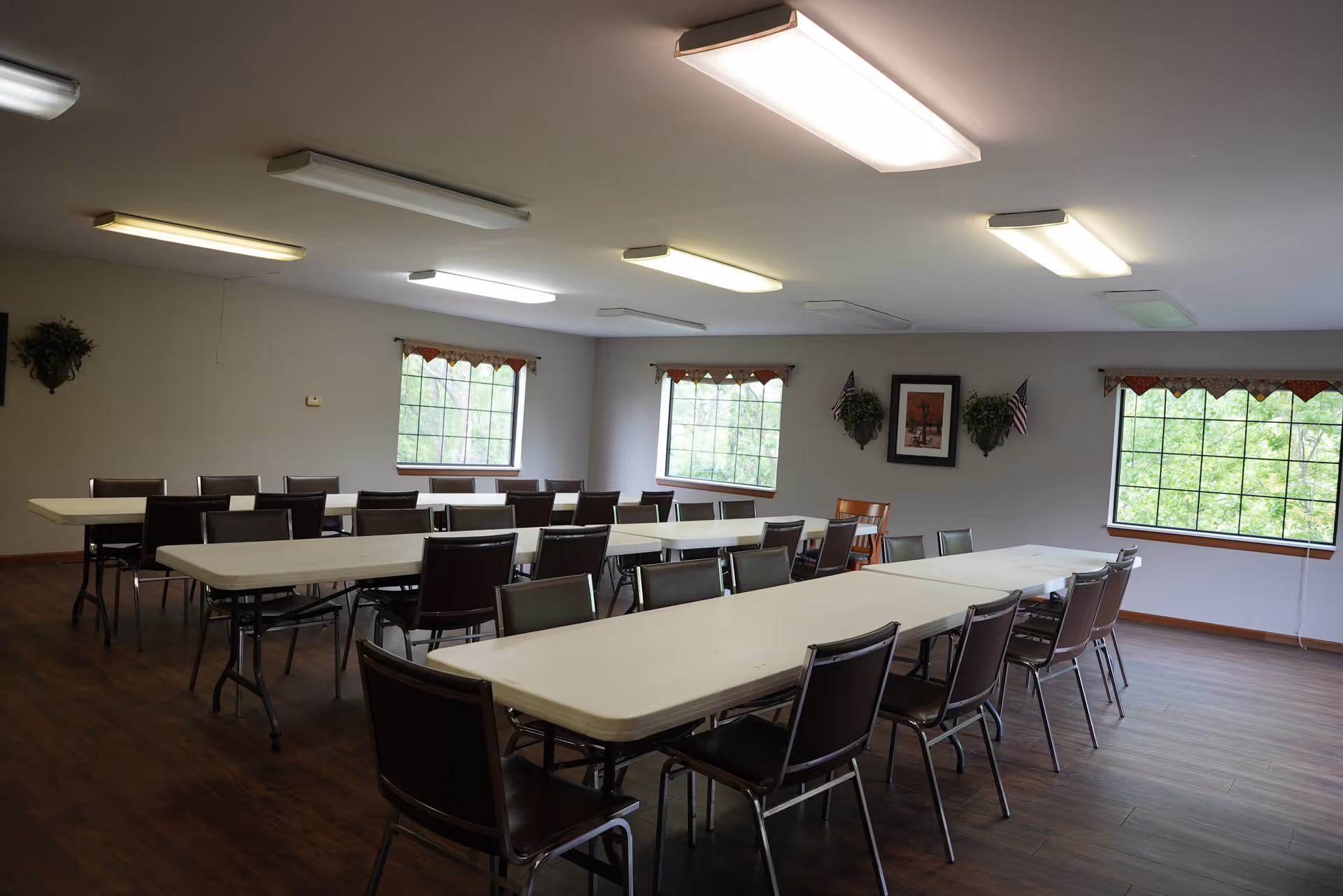 A large multipurpose dining/activity room with long folding tables, rows of chairs, windows and ceiling lights.