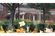 A garden area with colorful flowers in the foreground and a white gazebo surrounded by greenery and trees. A brick building is visible in the background.