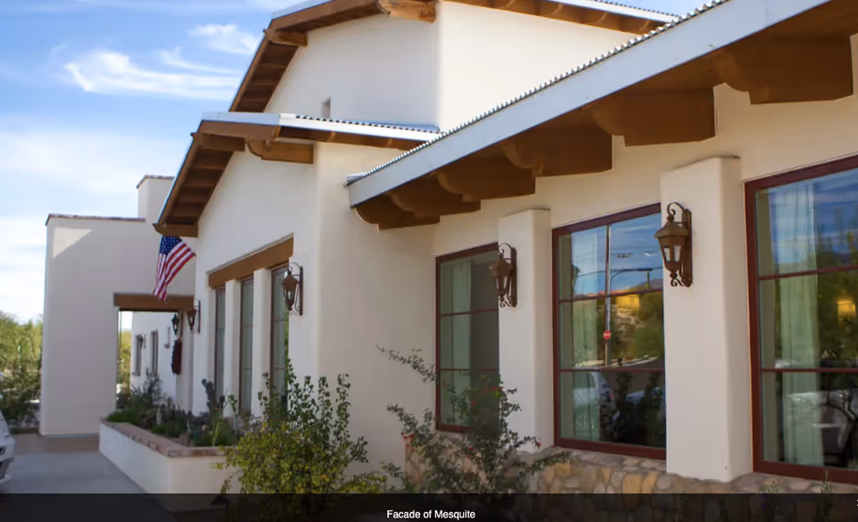 Exterior view of The Hacienda at the River building showing a beige facade with wooden beams under the roof, multiple windows with red frames, wall-mounted lantern-style lights, some greenery in front, and an American flag near the entrance.