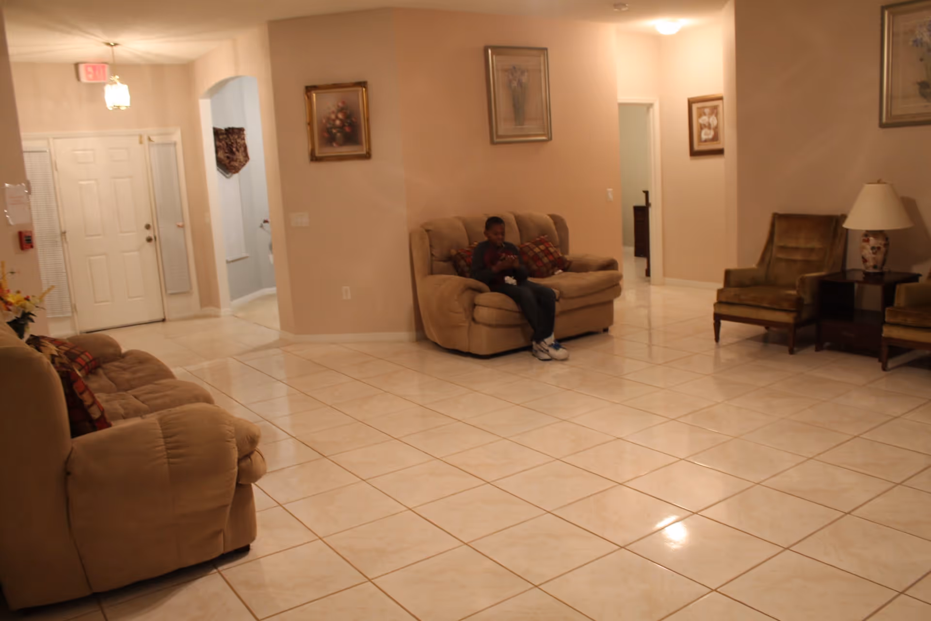 A spacious living room with beige tiled floors and light-colored walls. The room contains two beige sofas, one with a young boy sitting on it, and two brown armchairs with a small table and lamp between them. There are framed floral paintings on the walls and a white double door with windows covered by blinds near the entrance.