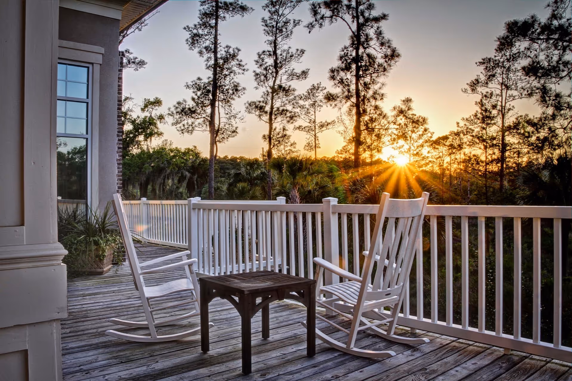 A wooden deck with two white rocking chairs and a small wooden table between them, overlooking a forested area with tall trees at sunset.