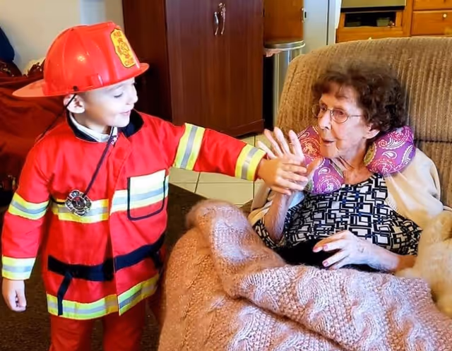 A young child dressed in a red firefighter costume is smiling and holding hands with an elderly woman who is sitting in a cushioned chair covered with a pink knitted blanket. The elderly woman is wearing glasses and a patterned top with a purple neck pillow. They are indoors in a cozy living space with wooden cabinets and tiled flooring visible in the background.