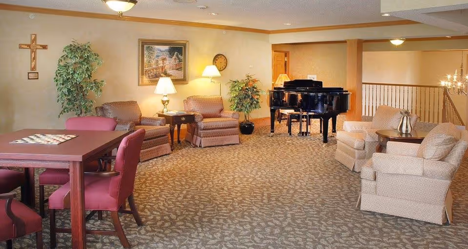 A spacious living room area with patterned carpet, several upholstered armchairs, a wooden table with four red cushioned chairs, a black grand piano, two potted plants, a wall clock, a framed painting, and a wooden cross on the wall.