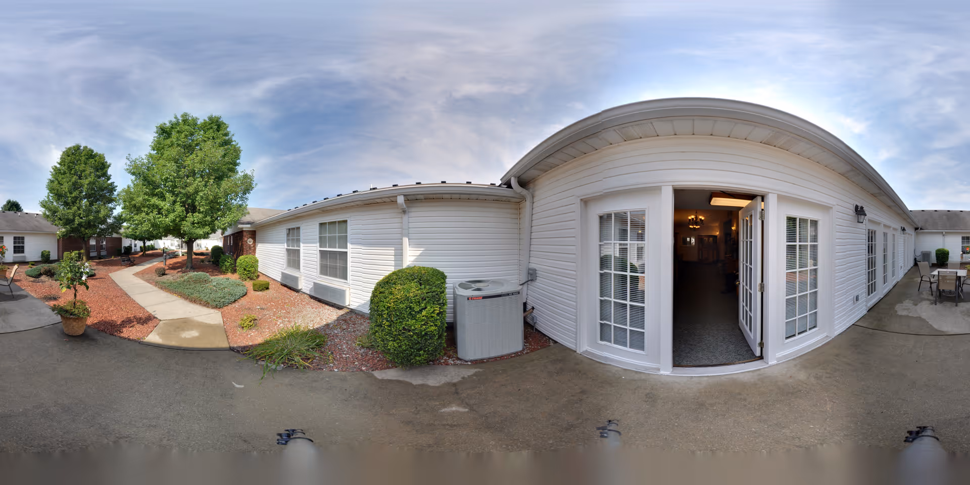 Outdoor courtyard area of a senior living facility with a paved walkway, landscaped garden beds with mulch, bushes, and trees. The building has white siding with multiple windows and French doors leading inside. Patio furniture is visible on the right side under a partly cloudy sky.