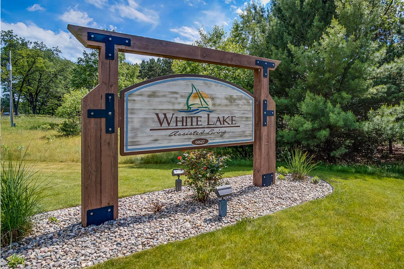 Wooden entrance sign that reads "White Lake Assisted Living" mounted on timber posts in a landscaped lawn with trees in the background.