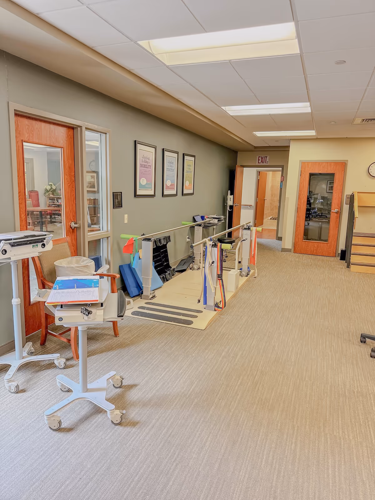Bright interior therapy area with parallel walking bars, medical carts on wheels, chairs, and framed posters along the wall.