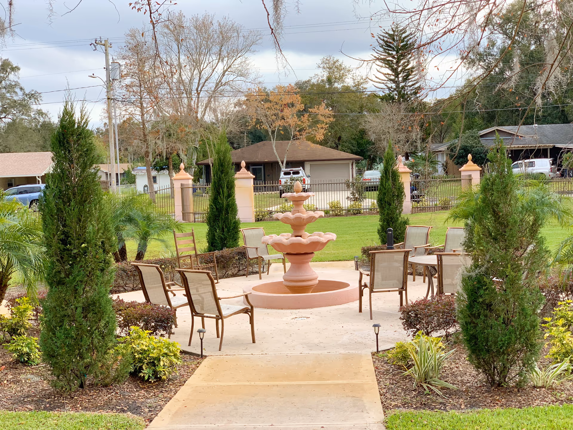 A landscaped outdoor courtyard centered on a three-tiered fountain with patio chairs and trees, with houses visible beyond.