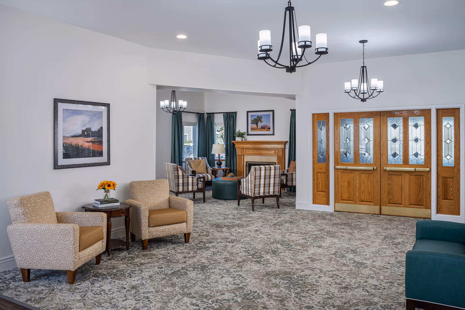 A spacious senior living facility common area with patterned carpet flooring, two beige armchairs with a small wooden side table holding a vase of sunflowers and books, a seating area with plaid armchairs around a wooden fireplace, green curtains on windows, and wooden double doors with decorative glass panels. Two black chandeliers with white lampshades hang from the ceiling.