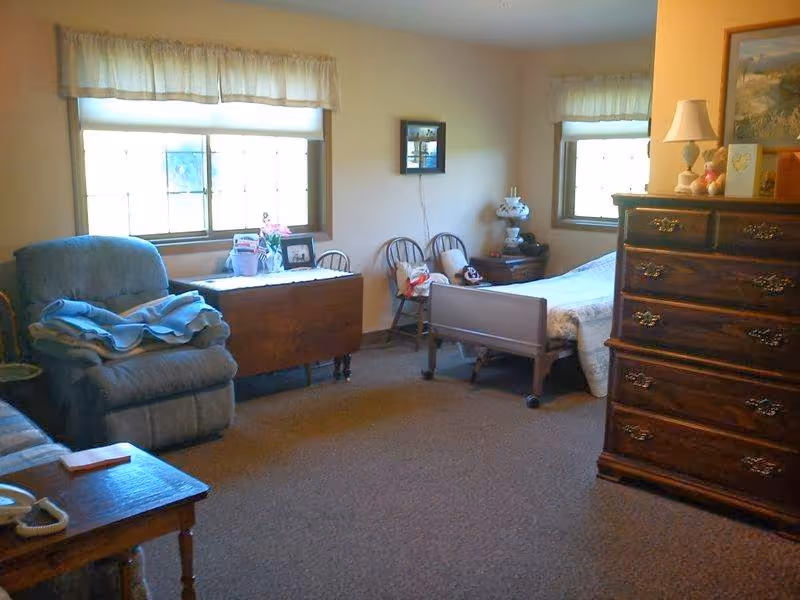 A cozy bedroom in an assisted living facility featuring a single bed with white bedding, a wooden dresser with a lamp and decorative items on top, two windows with light curtains, a comfortable recliner chair with a blanket, a wooden table with a telephone, and two wooden chairs against the wall.