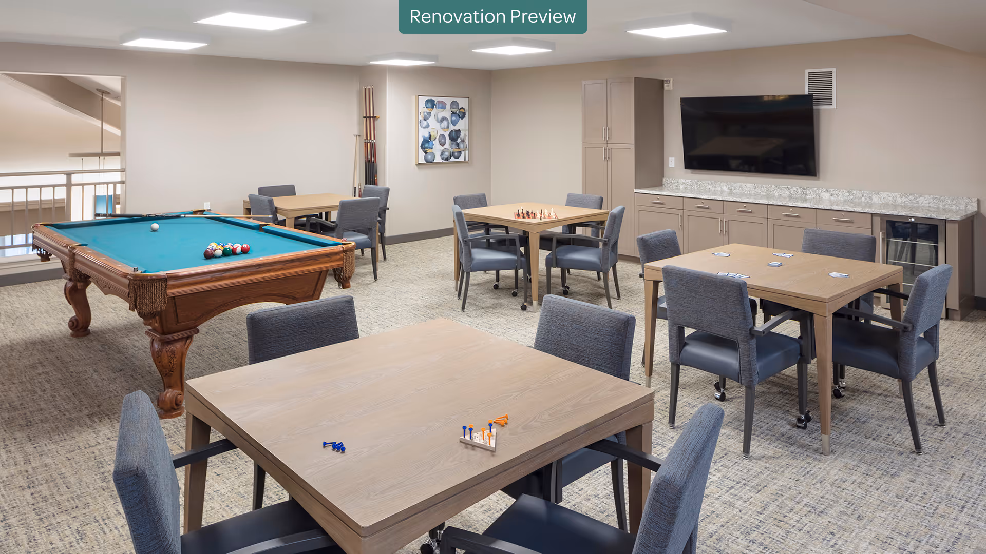A bright and spacious game room with several tables and chairs arranged for board games and card games. A pool table with balls and cues is positioned on the left side of the room. The back wall features a large flat-screen TV mounted above a long countertop with cabinets and a small refrigerator. The room has neutral-colored walls and carpeted flooring, with a modern and clean design.