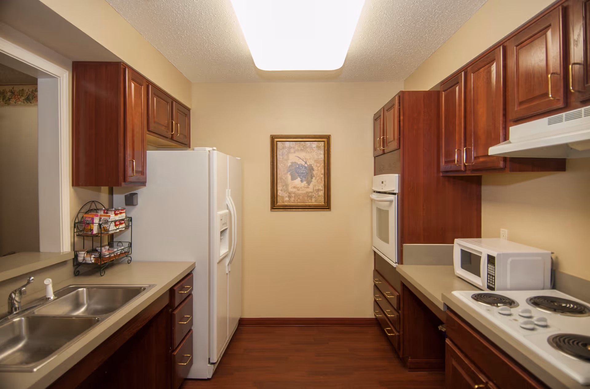 Galley kitchen with dark wood cabinets, a white refrigerator and oven, microwave and electric cooktop, and a double sink.