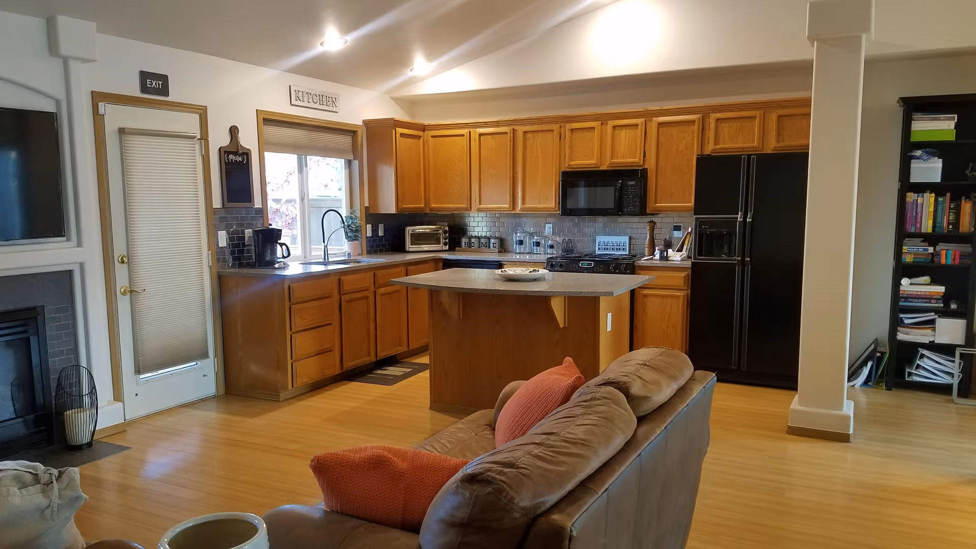 Interior view of a kitchen area with wooden cabinets, a black refrigerator, microwave, and stove. There is a kitchen island in the center with a decorative plate on top. To the left, there is a door with an exit sign above it and a coffee maker on the counter. In the foreground, part of a brown leather couch with orange pillows is visible. A bookshelf filled with books and items is seen on the right side.