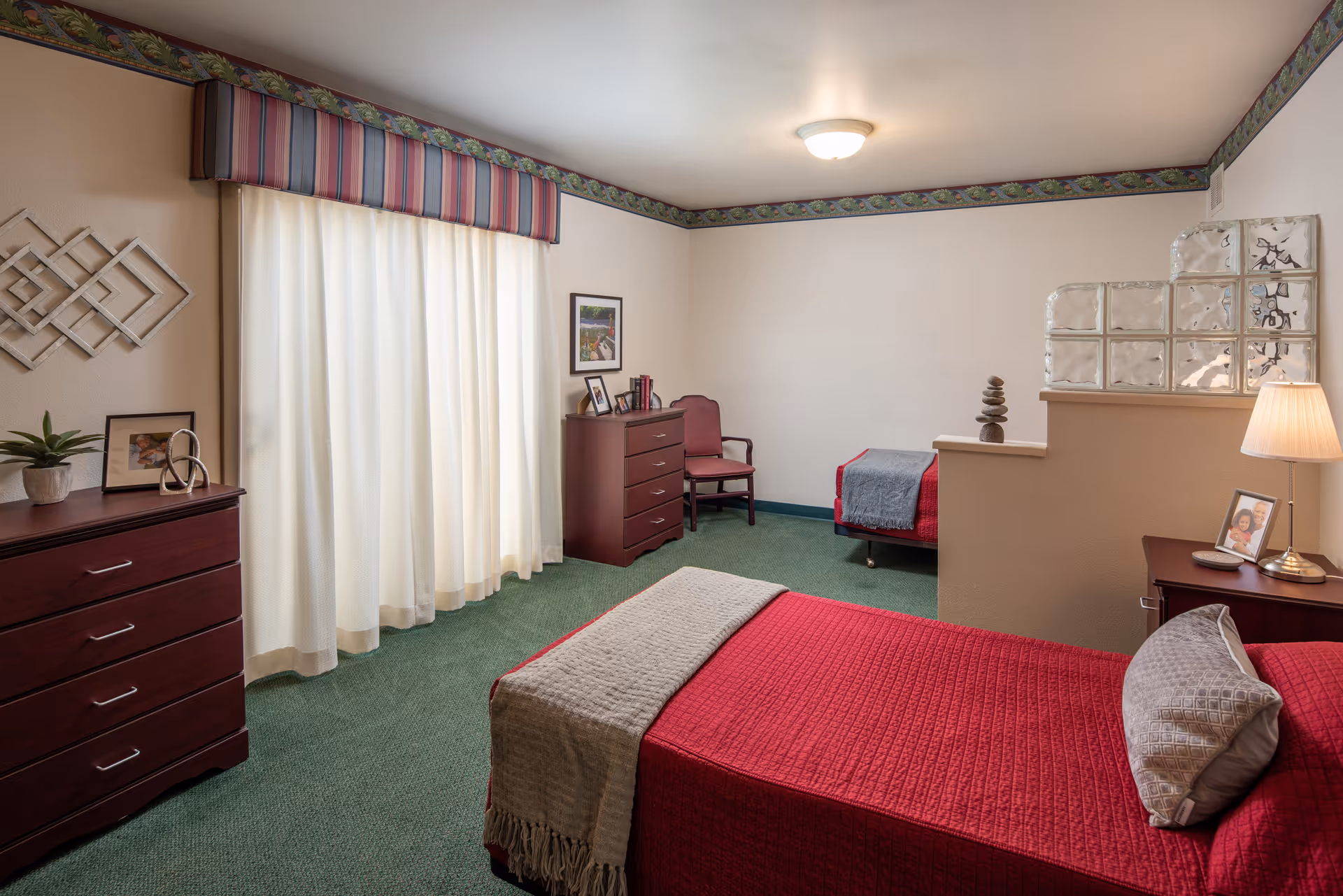 A cozy bedroom with two single beds covered in red bedspreads and gray throws. The room has green carpet, cream-colored walls with a floral border near the ceiling, and a window with white curtains and a striped valance. There are two wooden dressers, a wooden chair, a table lamp, framed photos, and decorative items on the furniture. A glass block partition partially separates the beds.