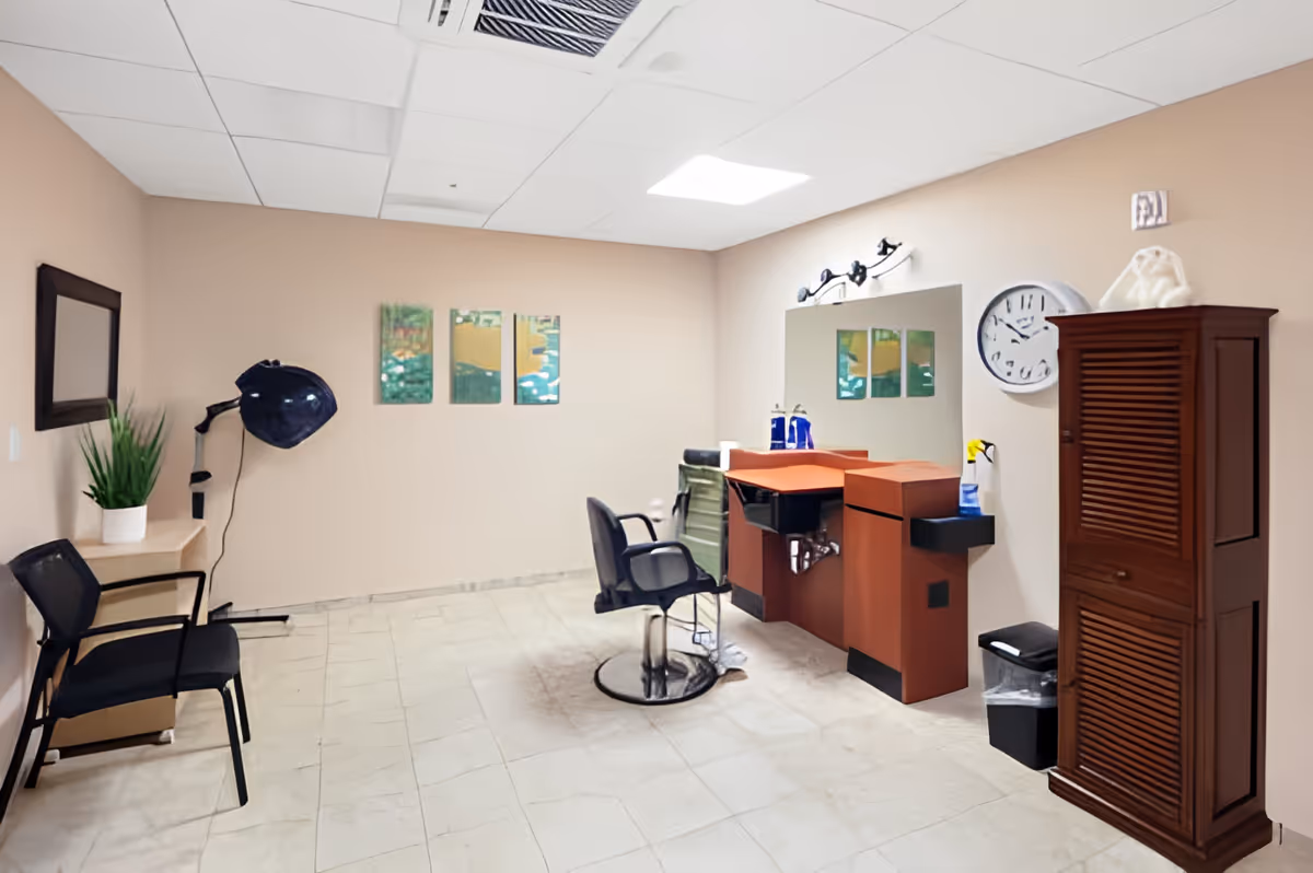 Interior of a salon or grooming room with a black salon chair in front of a wooden counter with a sink and mirror. There is a black hair dryer attached to the wall, a black chair next to a small table with a potted plant, a wall clock, and a tall wooden cabinet. The walls are beige and the floor is tiled.