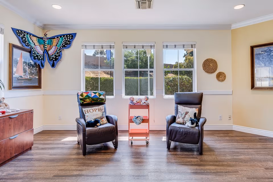 A bright room with two dark leather armchairs facing forward, each with decorative pillows and small stuffed animals. Between the chairs is a pink rolling cart with dolls on top. The room has three large windows letting in natural light, light yellow walls with white trim, wooden flooring, and wall decorations including a large colorful butterfly and framed paintings.