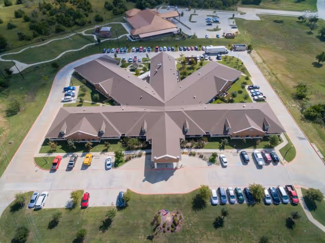 Aerial view of a large, star-shaped building with multiple wings, surrounded by parking lots and green lawns. Several cars are parked around the building, and there is a smaller building with a similar roof style nearby. The area is landscaped with trees and pathways.