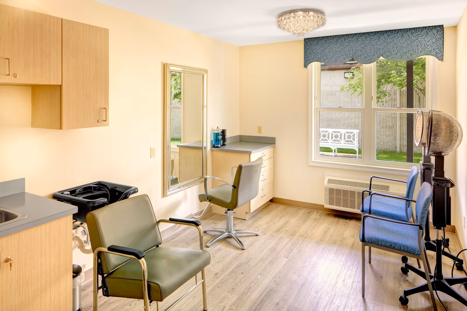 Bright hair-styling room with salon chairs, a mirror, sink, countertop and a hooded hair dryer by a window.