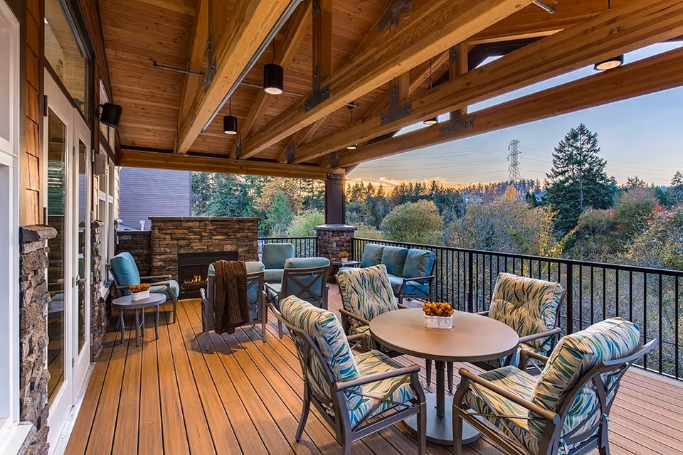 Covered outdoor patio with cushioned chairs around a round table, a stone fireplace, and a wooded view.