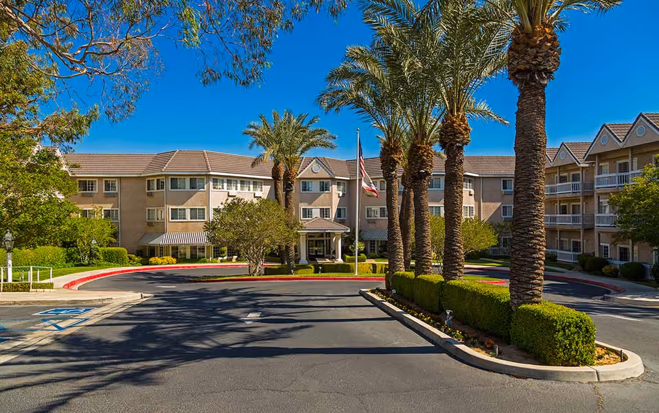 Front exterior view of Sterling Inn Assisted Living Community featuring a three-story building with balconies, palm trees lining the driveway, a flagpole with an American flag, and well-maintained landscaping under a clear blue sky.