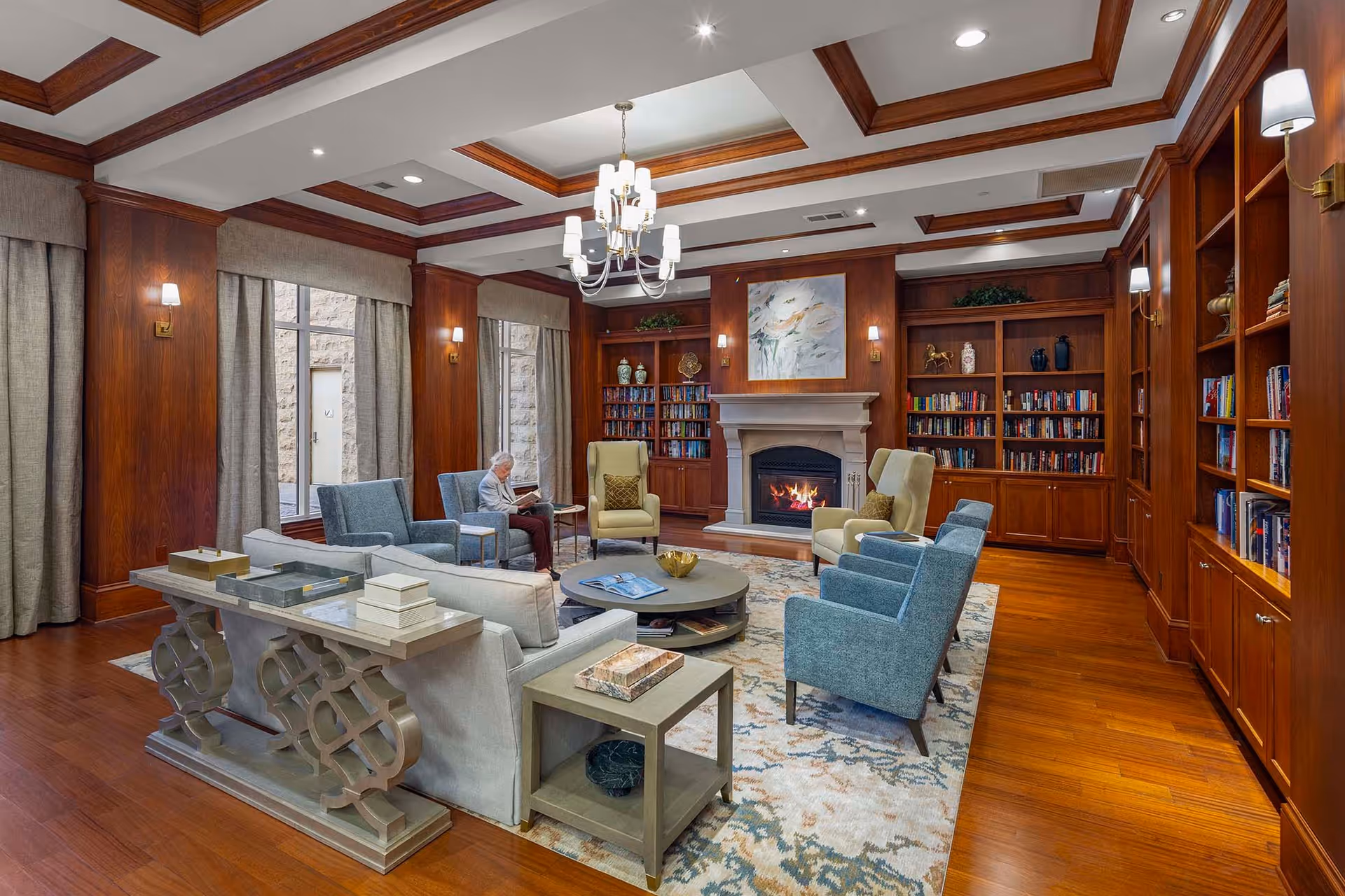 A cozy living room with wooden paneling and built-in bookshelves filled with books and decorative items. The room features a fireplace with a painting above it, a chandelier hanging from a coffered ceiling, and several upholstered chairs and sofas arranged around a round coffee table on a patterned rug. A person is seated near the window reading a book.