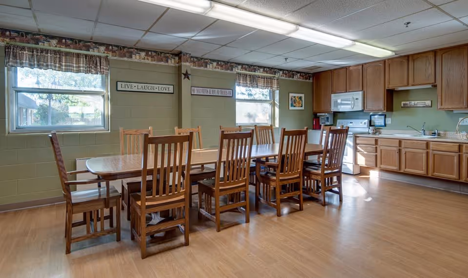 A communal dining area with a long wooden table surrounded by wooden chairs. The room has two windows with plaid valances, green walls decorated with framed signs and artwork, and a kitchen area with wooden cabinets, a microwave, stove, and sink. The floor is light wood, and the ceiling has fluorescent lighting.