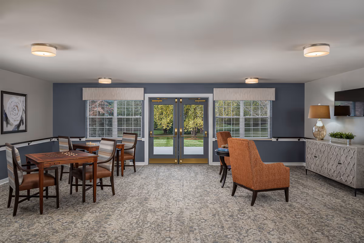 A well-lit common area in a senior living facility with two sets of tables and chairs arranged for games or socializing. The room features large windows with white blinds, double glass doors leading outside, a patterned carpet, and a sideboard with a lamp and a small plant. The walls are painted in a combination of blue and white, and there is a framed floral artwork on one wall.