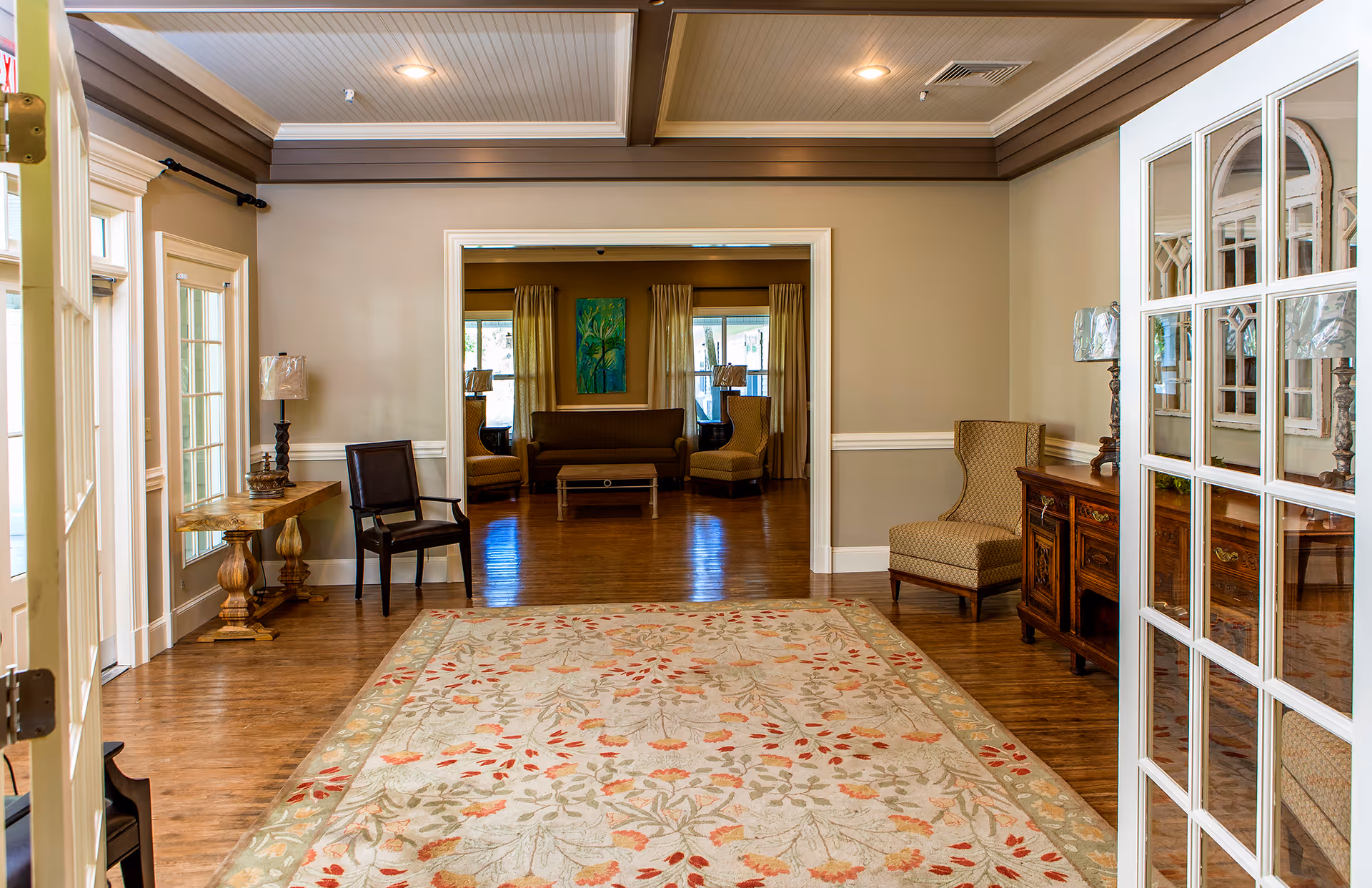 A spacious and well-lit interior common area in Fellowship Nashville Assisted Living Community featuring wooden floors, a large floral patterned area rug, several chairs, a wooden side table with a lamp, and a wooden cabinet with decorative lamps. The room opens into another seating area with a sofa, armchairs, and windows with curtains.