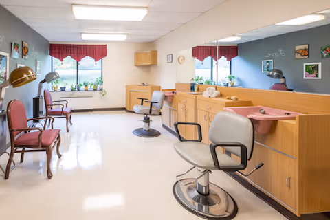 A bright salon room with styling chairs, hooded hairdryers, a pink wash sink, mirrors, and seating by windows with potted plants.