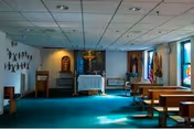 Interior view of a small chapel or prayer room with wooden pews arranged on both sides of a central aisle, a green carpeted floor, religious statues and crosses on the walls, and a small altar at the front with a crucifix and religious decorations.
