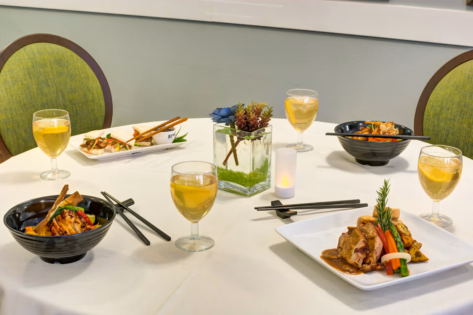 A dining table set with various dishes including grilled meat with vegetables, Asian-style dishes in black bowls, and spring rolls on a white rectangular plate. There are four glasses of iced tea with lemon, chopsticks, and a small decorative plant centerpiece on a white tablecloth. Two green cushioned chairs are partially visible.