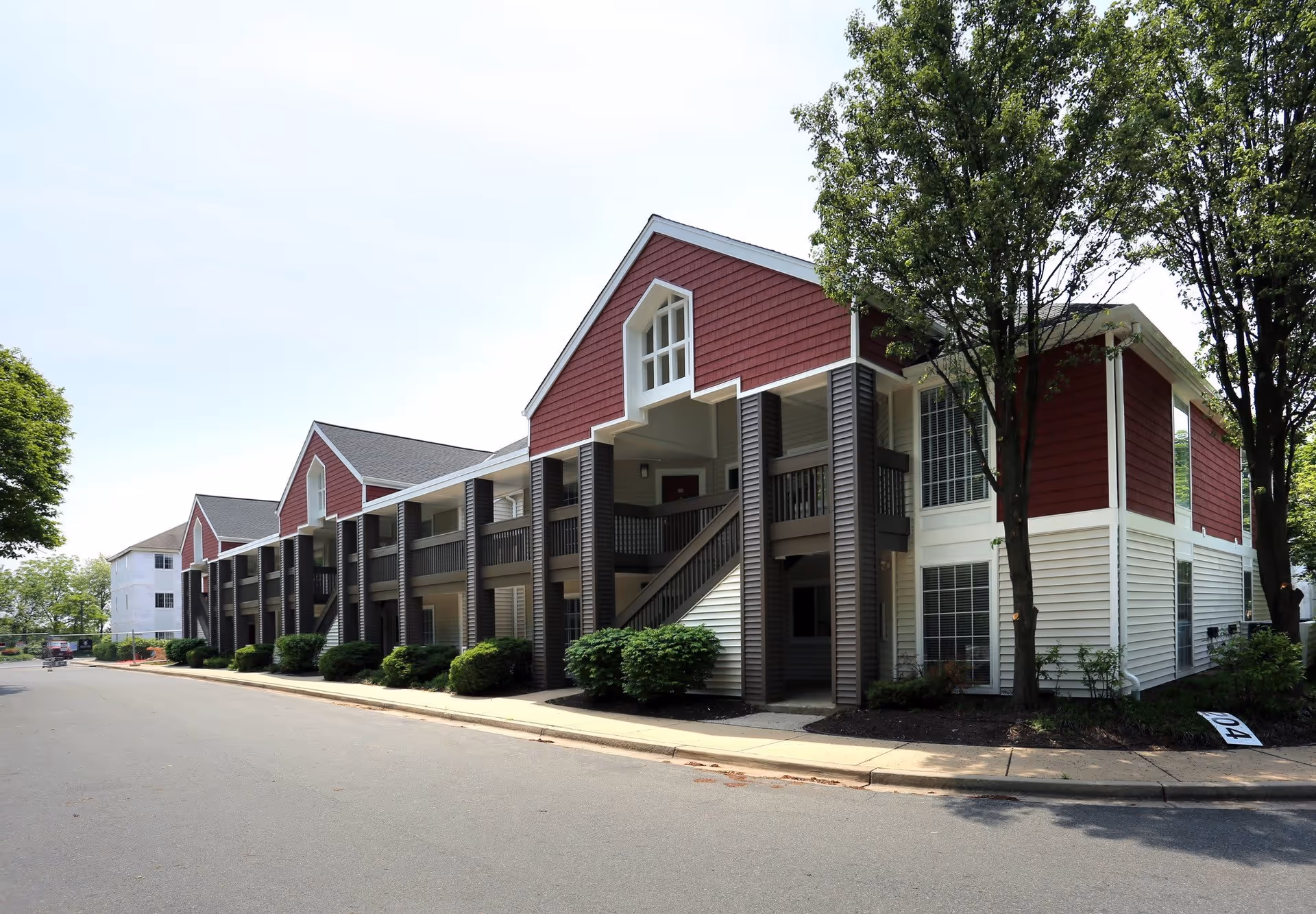 Exterior view of a senior living facility building with red and white siding, multiple windows, and an outdoor staircase. The building is surrounded by trees and bushes, with a paved road in front.