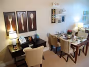 A dining area with a wooden table set for four people with plates, cups, and a flower centerpiece. Four beige upholstered chairs surround the table. To the left, there is a small desk with a lamp, a phone, and a chair. The wall behind the desk features three large framed pictures of a fork, knife, and spoon. Additional decorative wall art is visible above and beside the dining table. The room is softly lit by a table lamp on the desk and another lamp on a side table near the dining area.