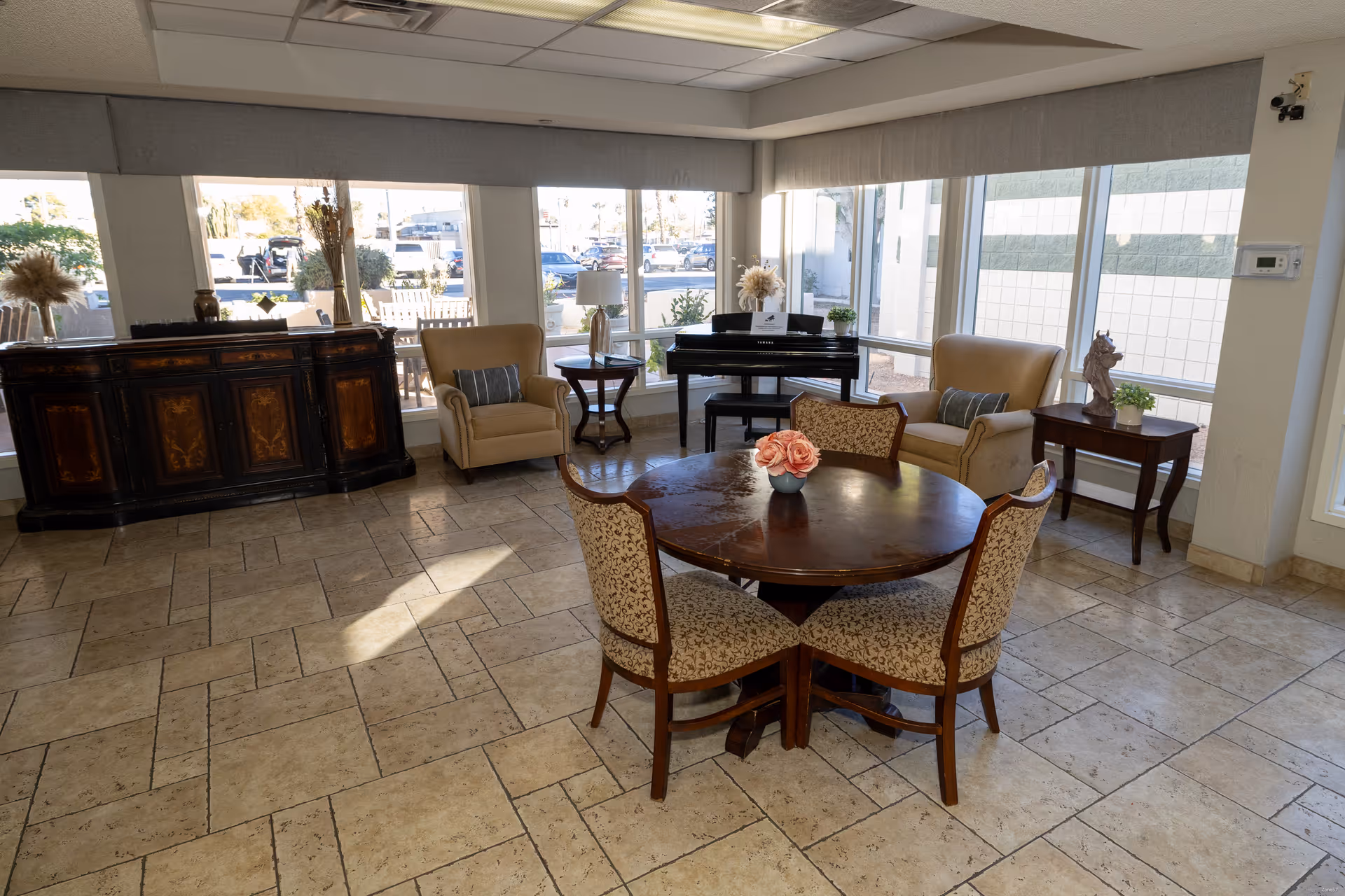 A bright room with large windows showing an outdoor parking area. Inside, there is a round wooden table with four upholstered chairs, a grand piano with a bench, two beige armchairs with pillows, and two small side tables with decorative items. A dark wooden cabinet is positioned against the wall on the left side.