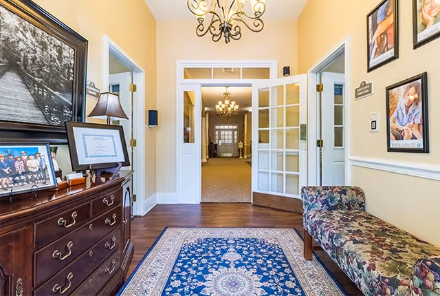 A well-lit interior hallway with yellow walls and wooden flooring. On the left side, there is a dark wooden dresser with framed photos and a lamp. On the right side, there is a floral upholstered bench beneath framed pictures on the wall. At the end of the hallway, double glass doors open into another room with carpeted flooring and chandeliers.