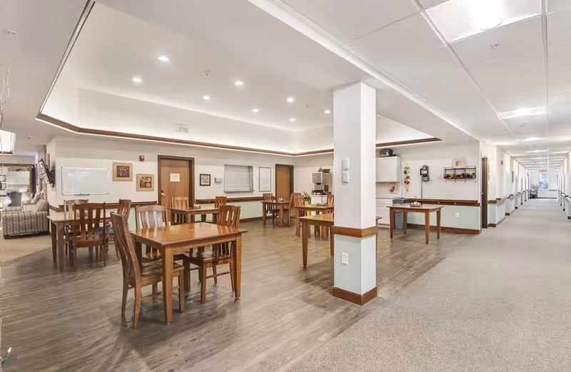 Interior view of a senior living facility common area with wooden tables and chairs arranged on a wood floor. The space is well-lit with recessed ceiling lights and has a long hallway with carpet extending to the right. Walls are decorated with framed pictures and a whiteboard, and there is a small kitchenette area visible in the background.