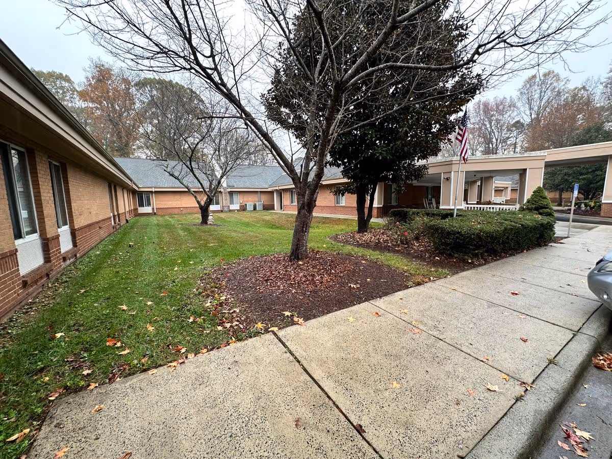 Front courtyard and covered entrance of a one-story brick senior living building with trees, a sidewalk, and an American flag.