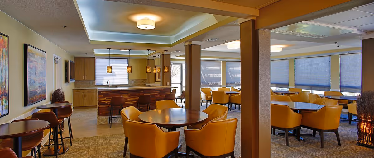 A spacious dining area in a senior living facility with multiple round tables surrounded by yellow upholstered chairs. The room features large windows with blinds, pendant lights hanging over a wooden counter, and modern ceiling lights. There are also high tables with bar stools along one wall and colorful artwork on the walls.