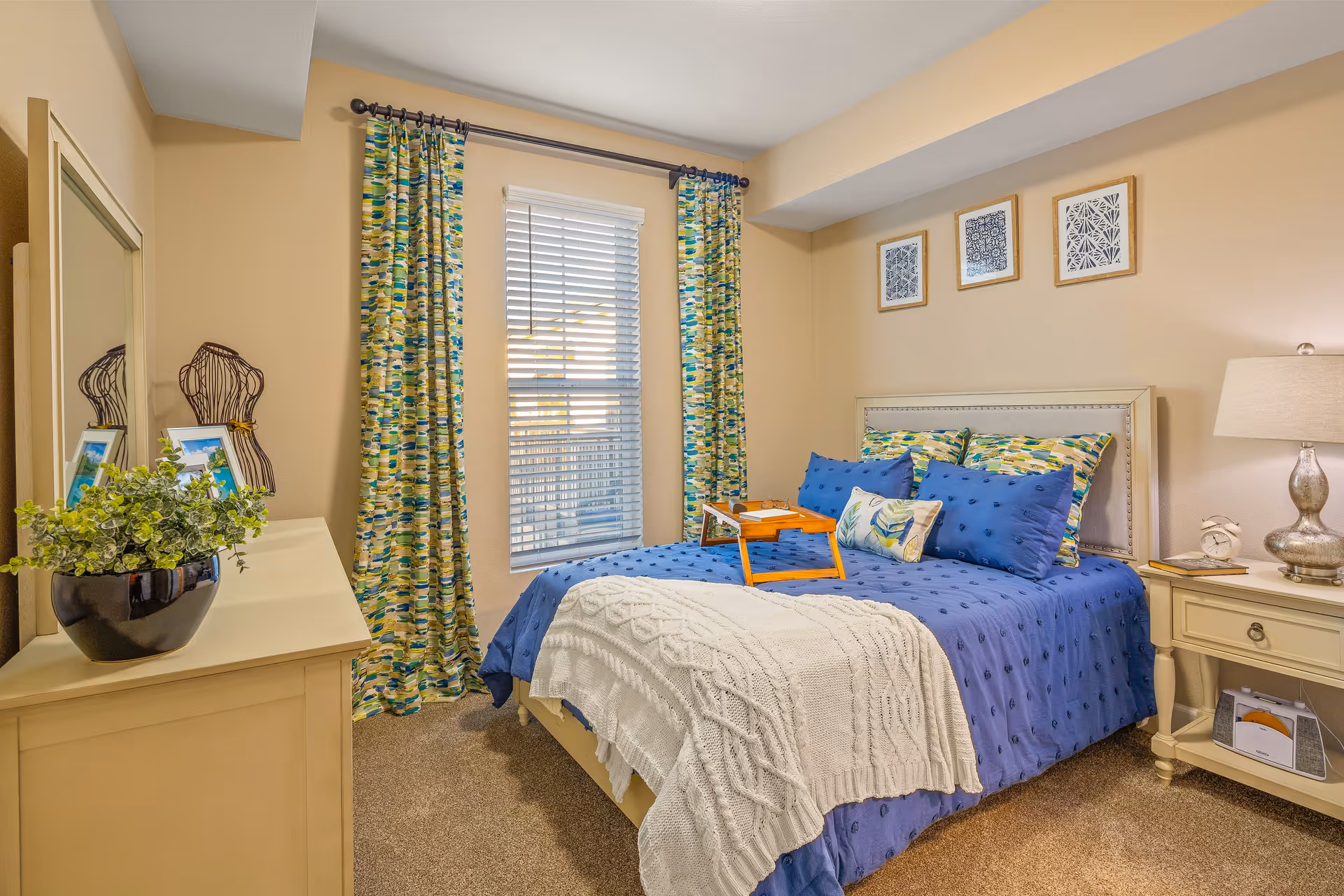 A cozy bedroom with a bed dressed in blue bedding and patterned pillows. A white knitted throw blanket is draped over the foot of the bed. There is a wooden tray on the bed. The room has beige walls with three framed artworks above the headboard. A window with colorful patterned curtains and white blinds is on one wall. A cream-colored dresser with a black pot plant and two framed photos is on the left side, and a matching nightstand with a lamp and clock is on the right side.
