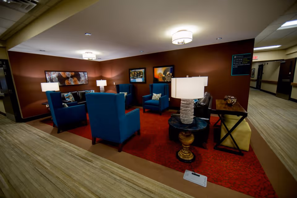 A cozy seating area in a senior living facility with four blue armchairs and a black leather couch arranged around a red patterned rug. The walls are painted brown and decorated with framed artwork. There are two table lamps on side tables and a standing lamp providing warm lighting. The area is adjacent to a hallway with light wood flooring and beige walls.