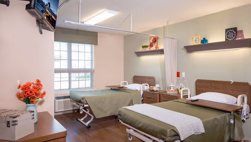 A bright and clean nursing facility room with two hospital beds covered in green blankets and white pillows. Each bed has a wooden headboard and an overbed table. There is a window with blinds letting in natural light, a wall-mounted TV in the corner, and a nightstand with decorative items between the beds. The room has wooden flooring and light-colored walls with shelves holding small decorative pieces.