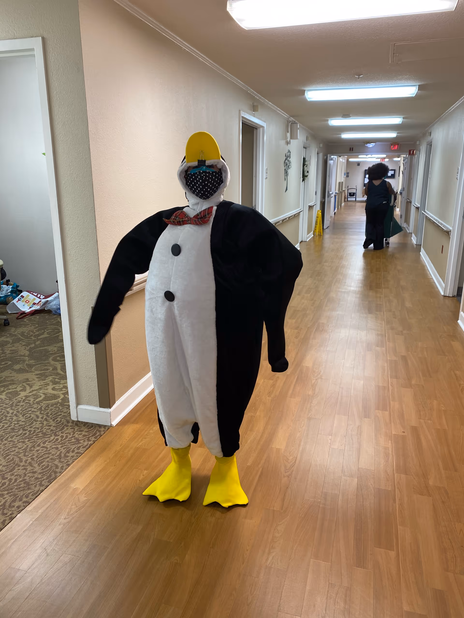 A person dressed in a penguin costume with a yellow beak and feet, black and white body, and a red bow tie stands in a hallway of a senior living facility. The person is also wearing a black face mask with white polka dots. The hallway has wooden flooring, beige walls, and several doors along the sides. Another person is visible walking away in the background.